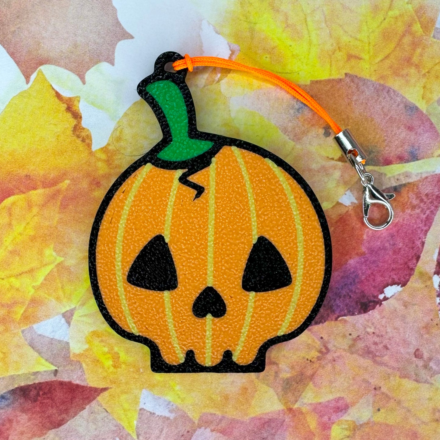 Pumpkin skull-shaped Tab Buddy  keychain with a green stem on a colorful leaf background