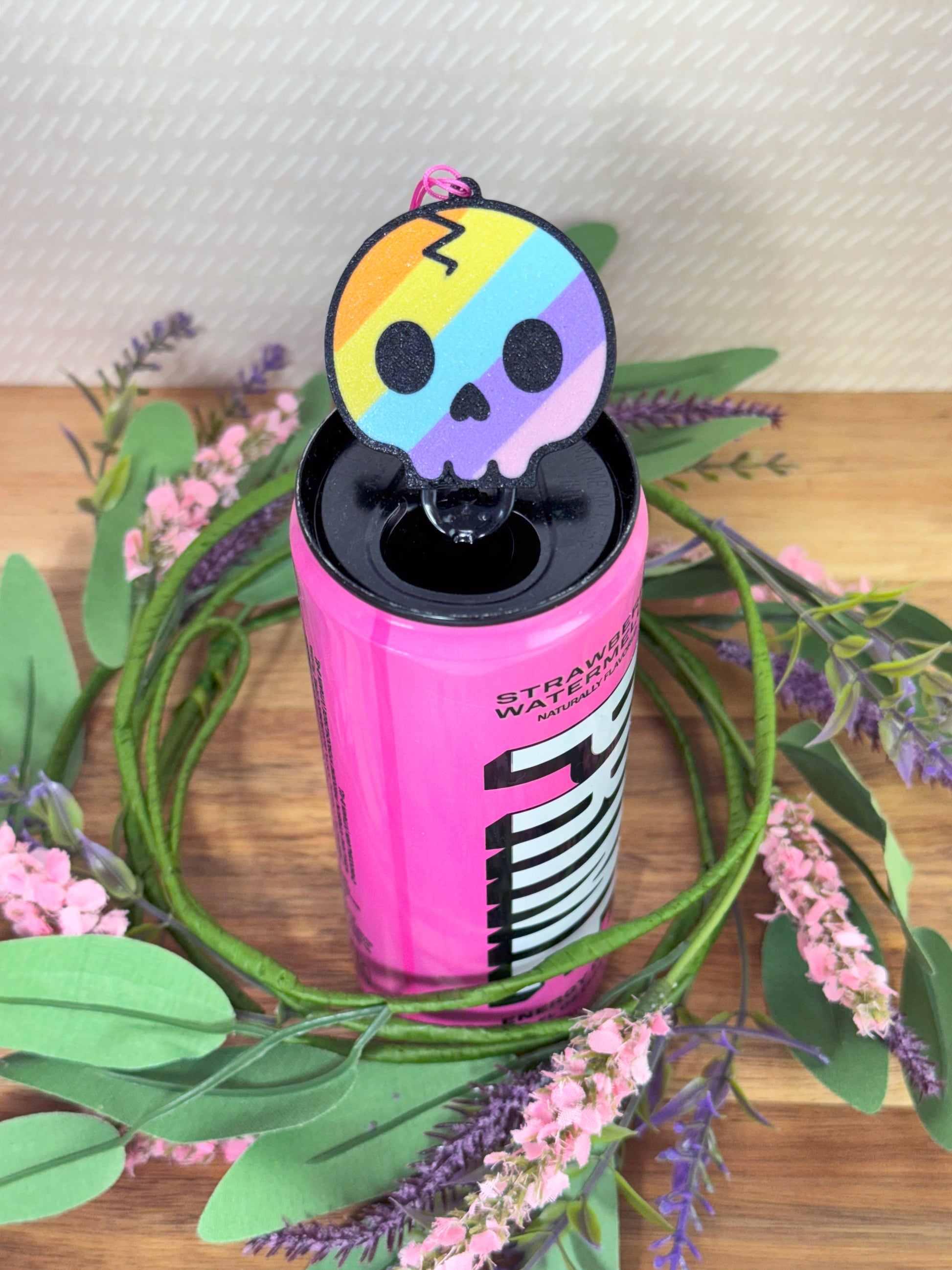 Pink energy drink can with a rainbow skull design Tab Buddy on top, surrounded by green leaves and purple flowers.
