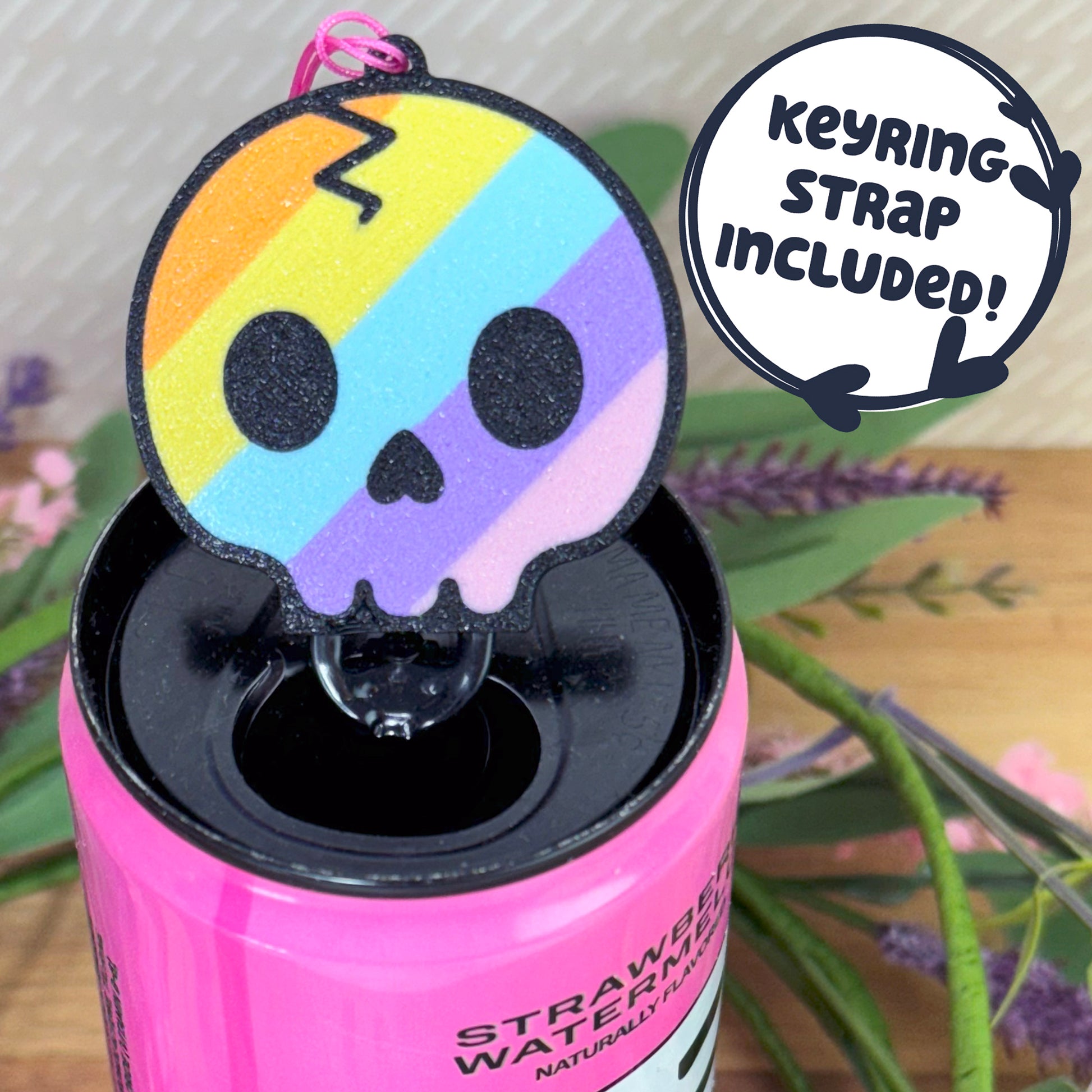 Pink energy drink can with a rainbow skull Tab Buddy with keyring strap on top, surrounded by plants.