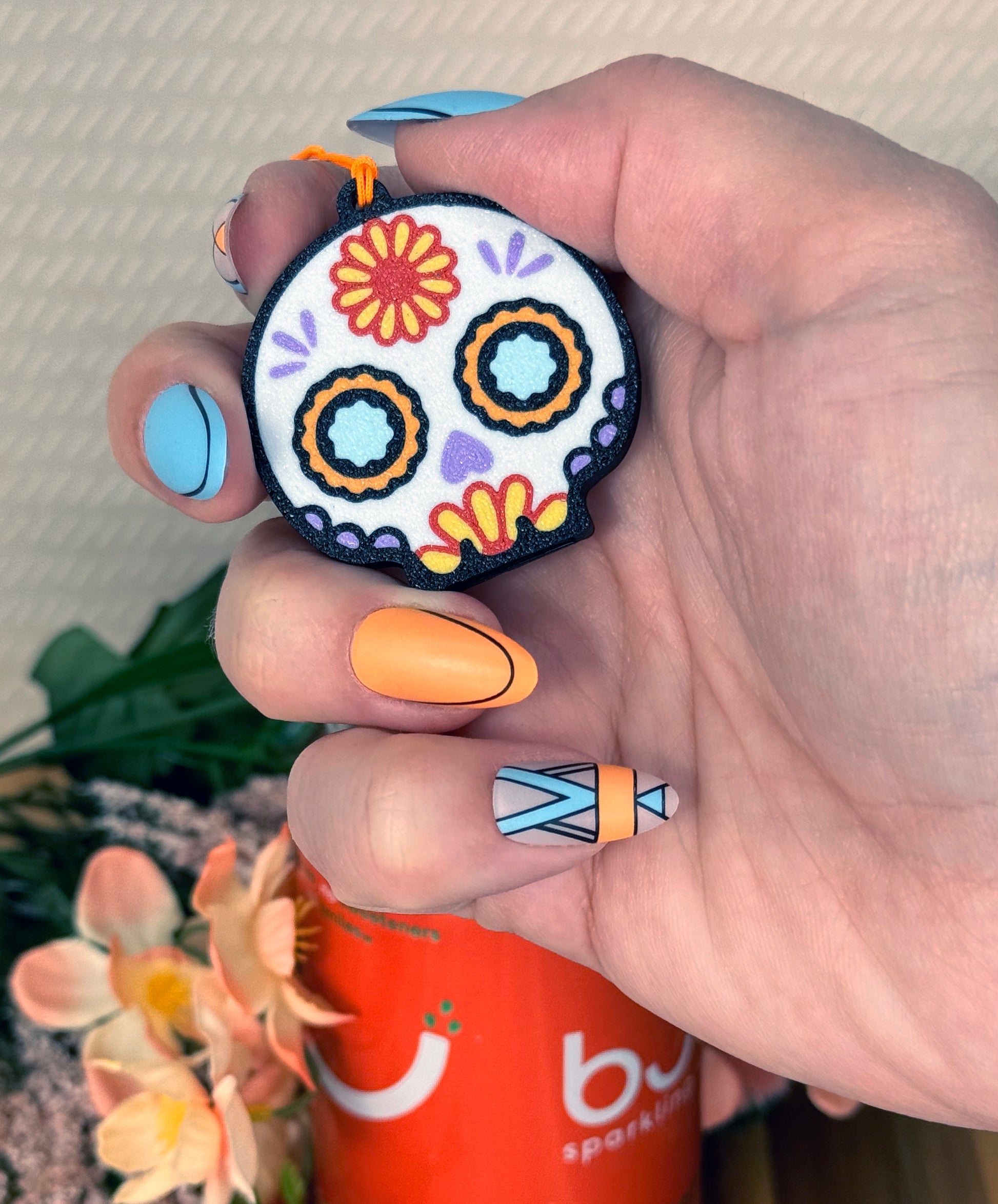 Hand with long styled nails holding a colorful sugar skull-shaped Tab Buddy  with a blurred background