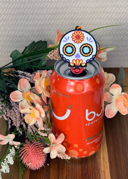 Orange can of sparkling water with a colorful sugar skull design Tab Buddy on top, surrounded by flowers.