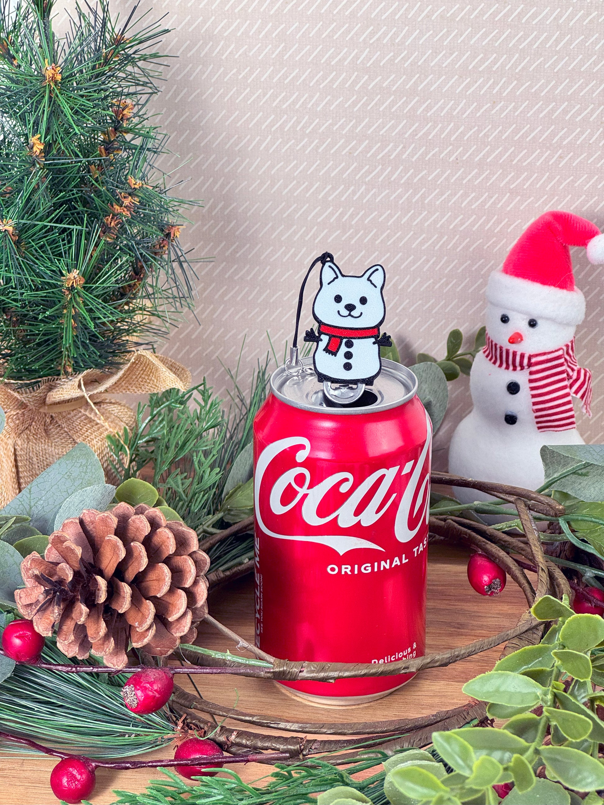 Coca-Cola can with a snowman dog design Tab Buddy on top, surrounded by Christmas decorations.
