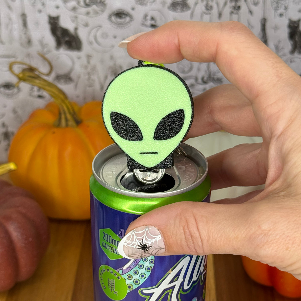 A hand holds a alien-shaped Soda Tab Buddy can opener on an energy drink can, with pumpkins and a patterned backdrop in the background.
