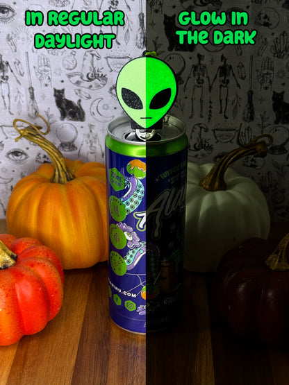 A split comparison of an alien head Soda Tab Buddy shown in regular daylight and glow-in-the-dark conditions. The center shows a split screen: the left half is in regular daylight and the alien head is a light shade of green, and the right half is dark with the same alien head Tab Buddy glowing bright green.