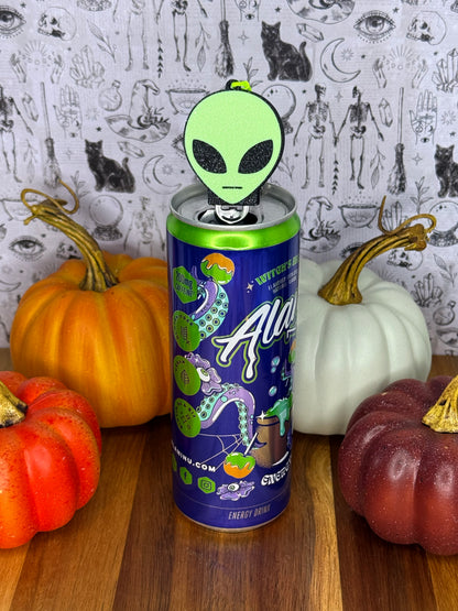A green alien head Soda Tab Buddy rests atop an energy drink can, styled on a wooden surface with orange, white, and red pumpkins against a black-and-white patterned backdrop.