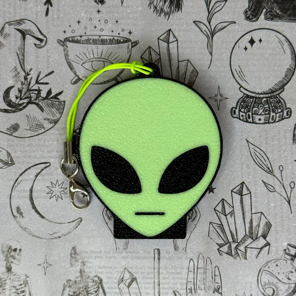 An alien-head Soda Tab Buddy with keychain strap rests on a patterned grey background with occult and crystal illustrations. The alien has a bright neon green face, black eyes, and a flat-line mouth.
