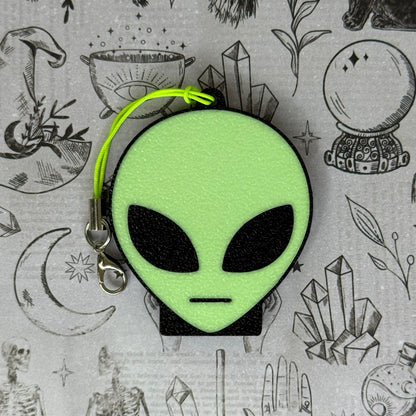An alien-head Soda Tab Buddy with keychain strap rests on a patterned grey background with occult and crystal illustrations. The alien has a bright neon green face, black eyes, and a flat-line mouth.
