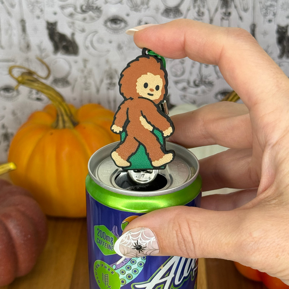 A Bigfoot-shaped Soda Tab Buddy with brown and tan fur and cute facial features rests atop the tab of an energy drink after opening it. A hand is atop the Tab Buddy after openinig the can. The can rests on a wooden surface with colorful pumpkins and a black-and-white wallpaper in the background.