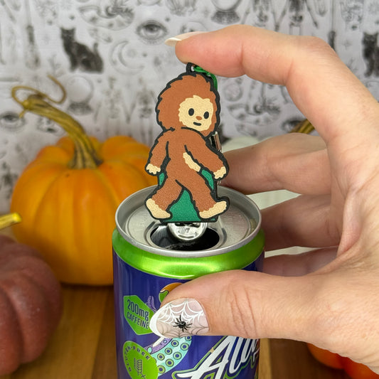 A Bigfoot-shaped Soda Tab Buddy with brown and tan fur and cute facial features rests atop the tab of an energy drink after opening it. A hand is atop the Tab Buddy after openinig the can. The can rests on a wooden surface with colorful pumpkins and a black-and-white wallpaper in the background.