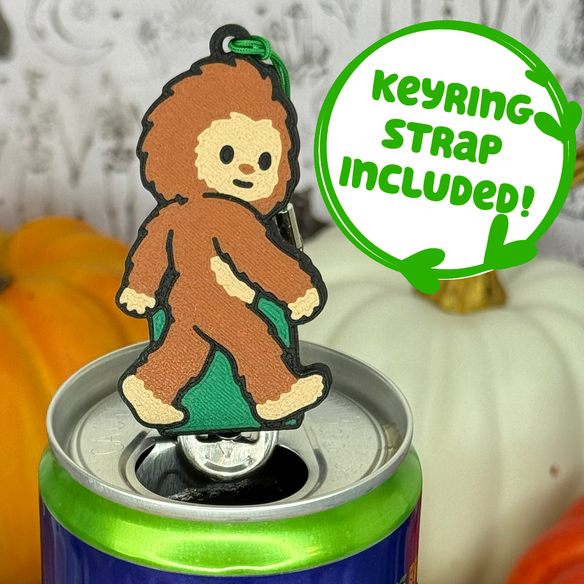 A Bigfoot-shaped Soda Tab Buddy with brown and tan fur and cute facial features is shown resting atop an open canned drink. A text bubble reads, 'Keyring Strap Included!' and the background includes pumpkins and a blurred indoor setting.