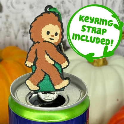 A Bigfoot-shaped Soda Tab Buddy with brown and tan fur and cute facial features is shown resting atop an open canned drink. A text bubble reads, 'Keyring Strap Included!' and the background includes pumpkins and a blurred indoor setting.
