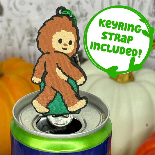 A Bigfoot-shaped Soda Tab Buddy with brown and tan fur and cute facial features is shown resting atop an open canned drink. A text bubble reads, 'Keyring Strap Included!' and the background includes pumpkins and a blurred indoor setting.
