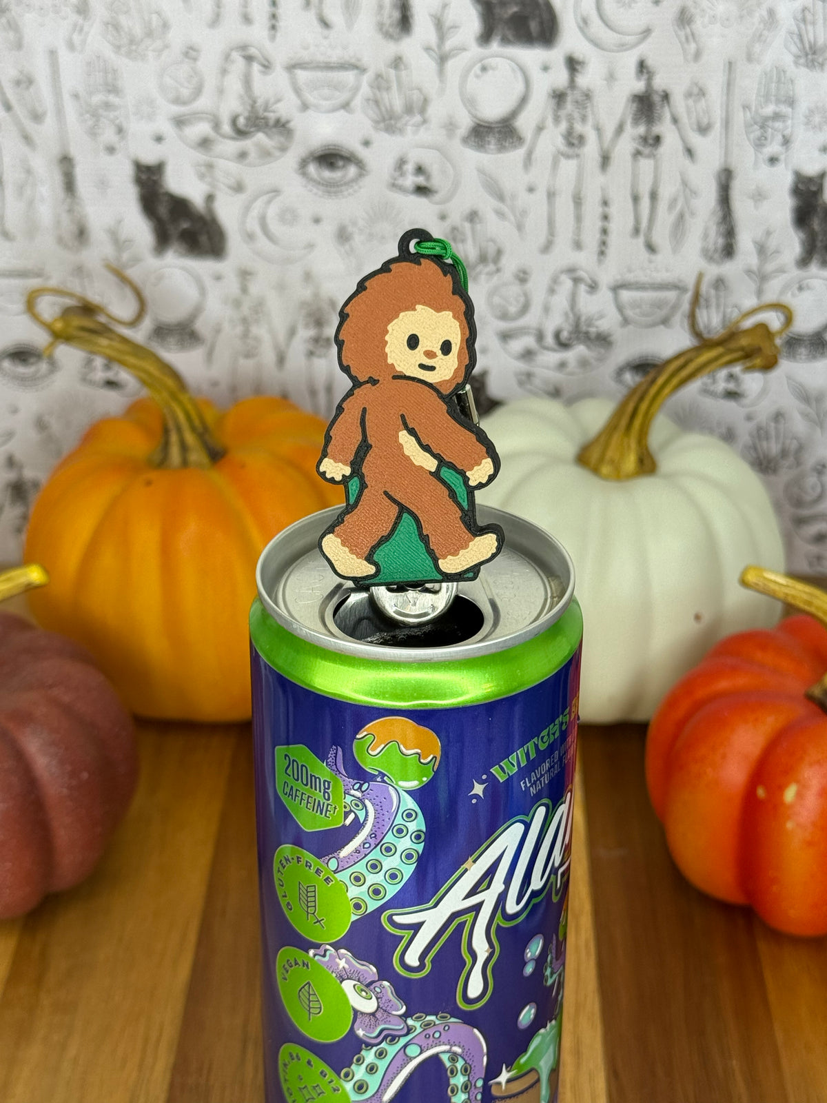 A Bigfoot-shaped Soda Tab Buddy with brown and tan fur and cute facial features rests atop the tab of an energy drink after opening it. The can rests on a wooden surface with colorful pumpkins and a black-and-white wallpaper in the background.