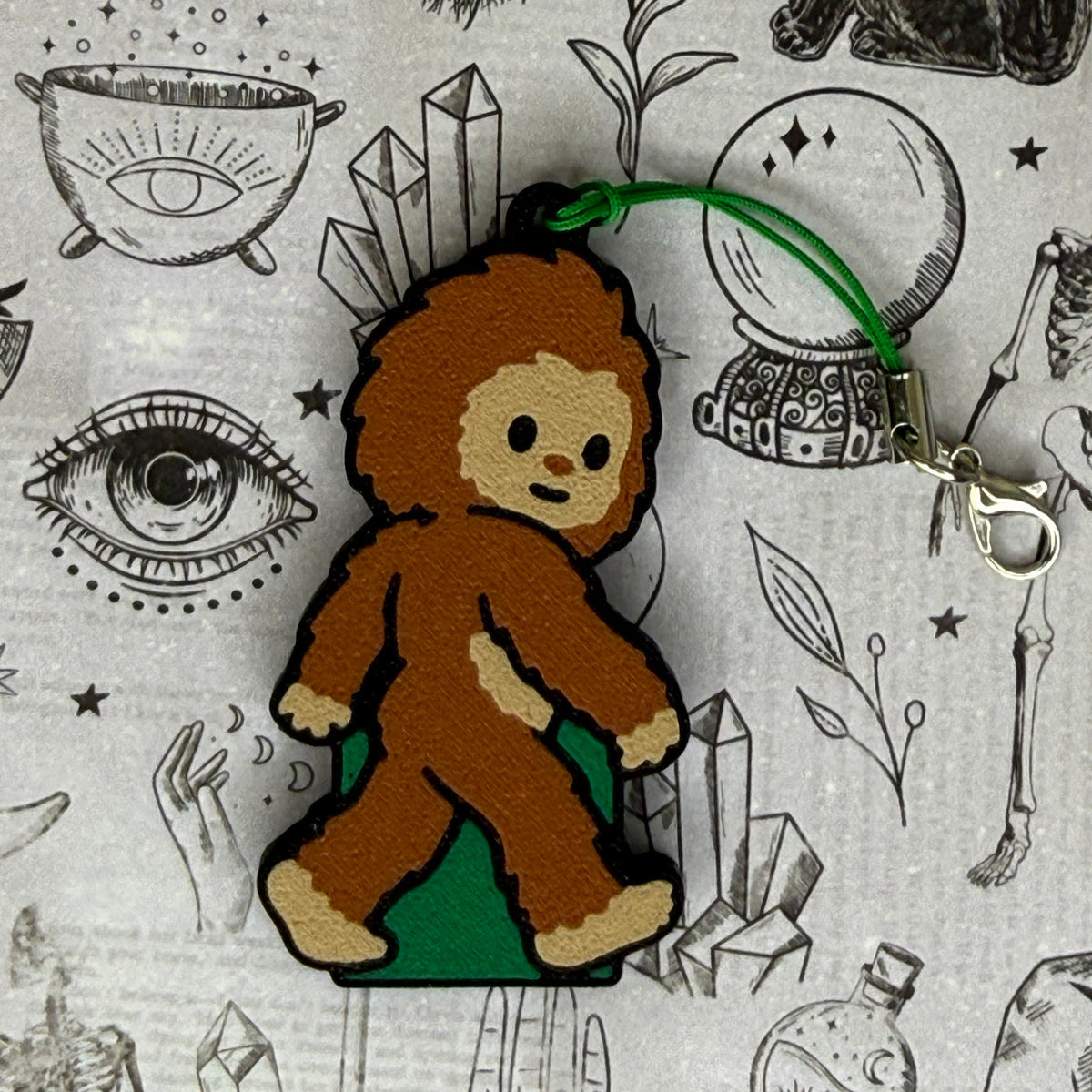 A Bigfoot-shaped Soda Tab Buddy with brown and tan fur and cute facial features and a green keyring strap rests atop a black-and-white spooky-themed illustration.