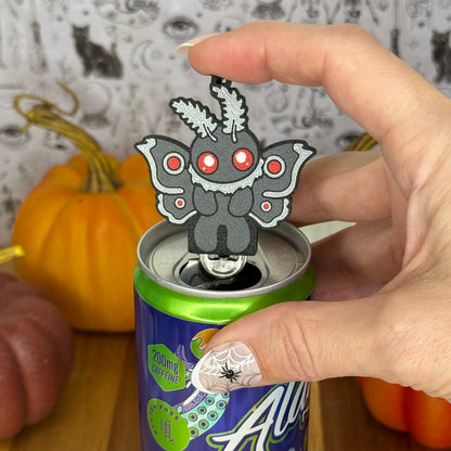 A Mothman-shaped Soda Tab Buddy with grey and silver wings and red eyes rests atop the tab of an energy drink after opening it. A hand rests on the Tab Buddy after just using it to open the can. The can rests on a wooden surface with colorful pumpkins and a black-and-white wallpaper in the background.