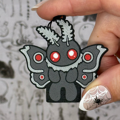 A Mothman-shaped Soda Tab Buddy with grey and silver wings, body, antennae and tufts is held in a hand with a light grey blurred wallpaper in the background.
