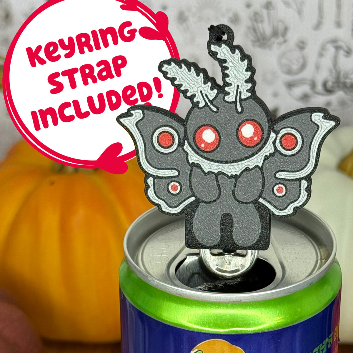A mothman-shaped Soda Tab Buddy with red eyes and patterned wings is shown resting atop an open canned drink. A text bubble reads, 'Keyring Strap Included!' and the background includes pumpkins and a blurred indoor setting.