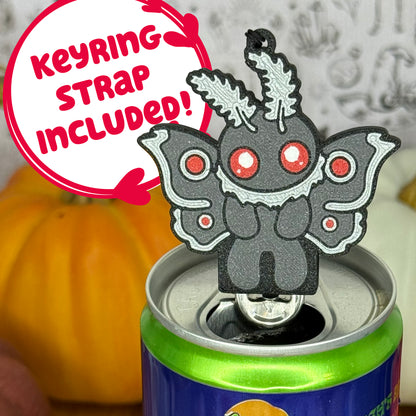 A mothman-shaped Soda Tab Buddy with red eyes and patterned wings is shown resting atop an open canned drink. A text bubble reads, 'Keyring Strap Included!' and the background includes pumpkins and a blurred indoor setting.