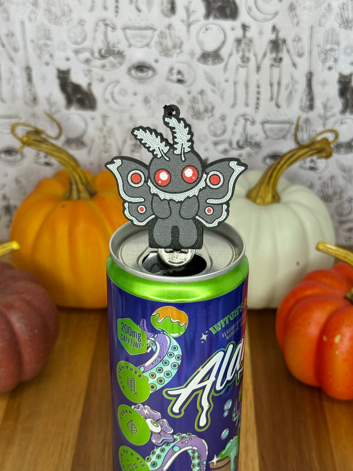 A Mothman-shaped Soda Tab Buddy with grey and silver wings and red eyes rests atop the tab of an energy drink after opening it. The can rests on a wooden surface with colorful pumpkins and a black-and-white wallpaper in the background.