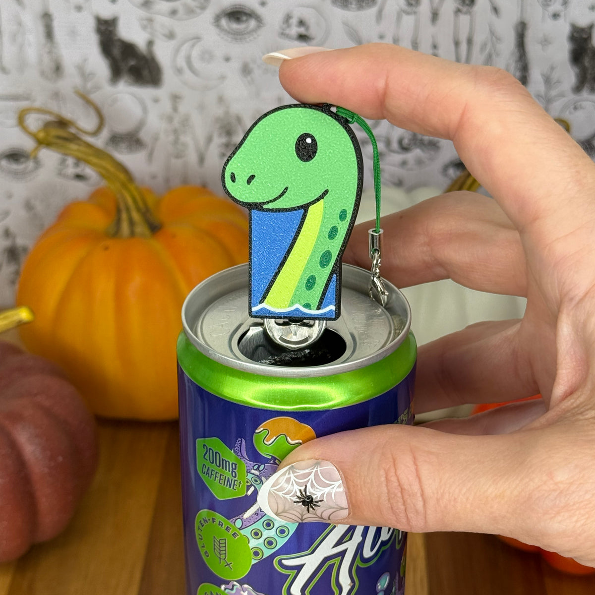 A Lock Ness Monster-shaped Soda Tab Buddy with green spots, cute facial features and a neck rising out of water rests atop the tab of an energy drink after opening it. A hand rests on the Tab Buddy after just using it to open the can. The can rests on a wooden surface with colorful pumpkins and a black-and-white wallpaper in the background.