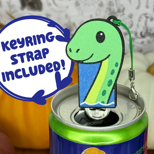 A Lock Ness Monster-shaped Soda Tab Buddy with cute facial features and a neck extending out of the water is shown resting atop an open canned drink. A text bubble reads, 'Keyring Strap Included!' and the background includes pumpkins and a blurred indoor setting.