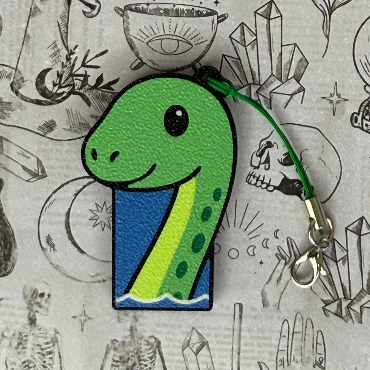 a cute green Loch Ness Monster Soda Tab Buddy rests atop a patterned background with spooky-themed illustrations. The Nessie is various shades of green with cute facial features, and appears to be rising out of a body of water.