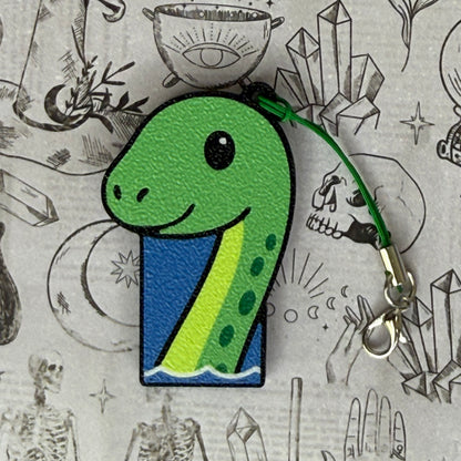 a cute green Loch Ness Monster Soda Tab Buddy rests atop a patterned background with spooky-themed illustrations. The Nessie is various shades of green with cute facial features, and appears to be rising out of a body of water.