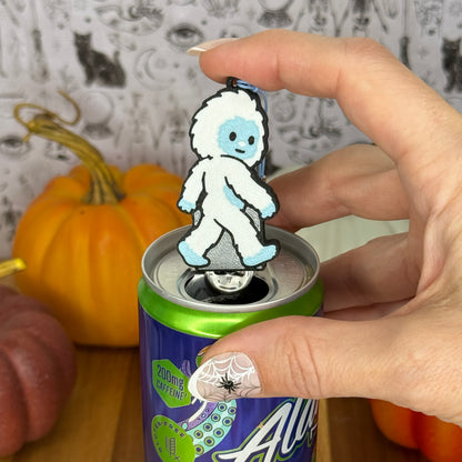 A Yeti-shaped Soda Tab Buddy with white and blue fur and cute facial features rests atop the tab of an energy drink after opening it. A hand rests on the Tab Buddy after just using it to open the can. The can rests on a wooden surface with colorful pumpkins and a black-and-white wallpaper in the background.