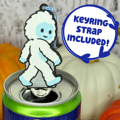 A Yeti-shaped Soda Tab Buddy with white and blue fur and cute facial features is shown resting atop an open canned drink. A text bubble reads, 'Keyring Strap Included!' and the background includes pumpkins and a blurred indoor setting.
