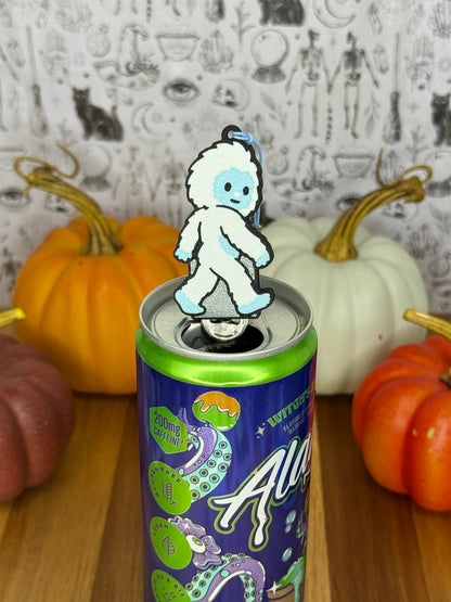 A Yeti-shaped Soda Tab Buddy with white and blue fur and cute facial features rests atop the tab of an energy drink after opening it. The can rests on a wooden surface with colorful pumpkins and a black-and-white wallpaper in the background.
