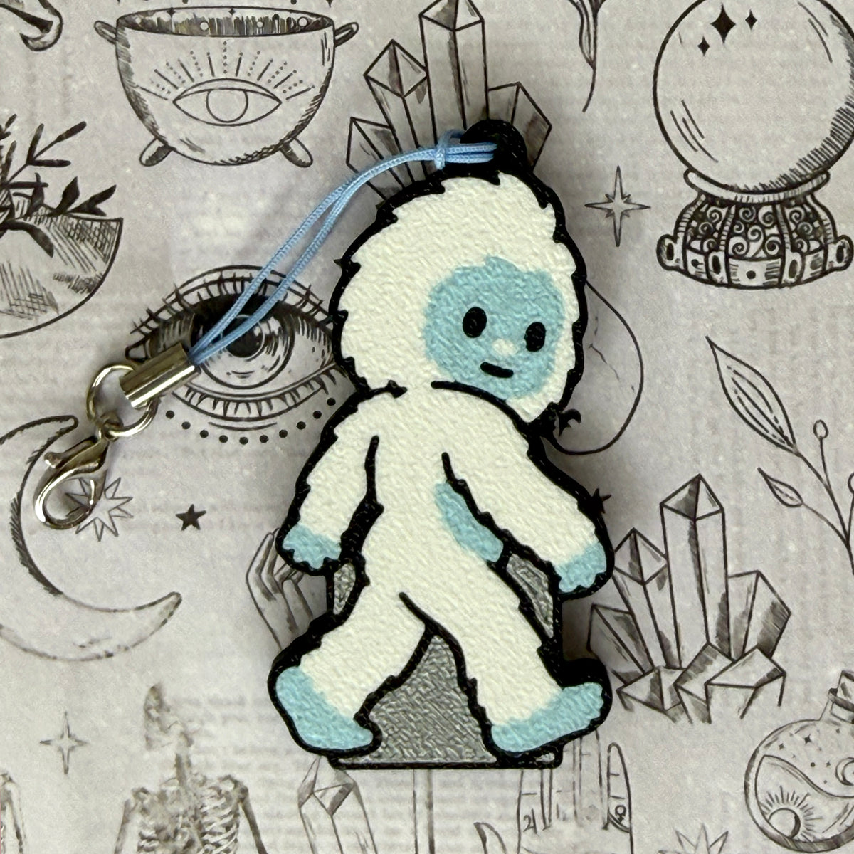 A Yeti-shaped Soda Tab Buddy with white and blue fur and cute facial features rests atop a black-and-white spooky-themed illustration.