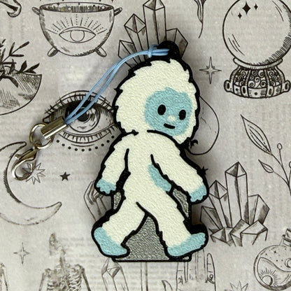 A Yeti-shaped Soda Tab Buddy with white and blue fur and cute facial features rests atop a black-and-white spooky-themed illustration.