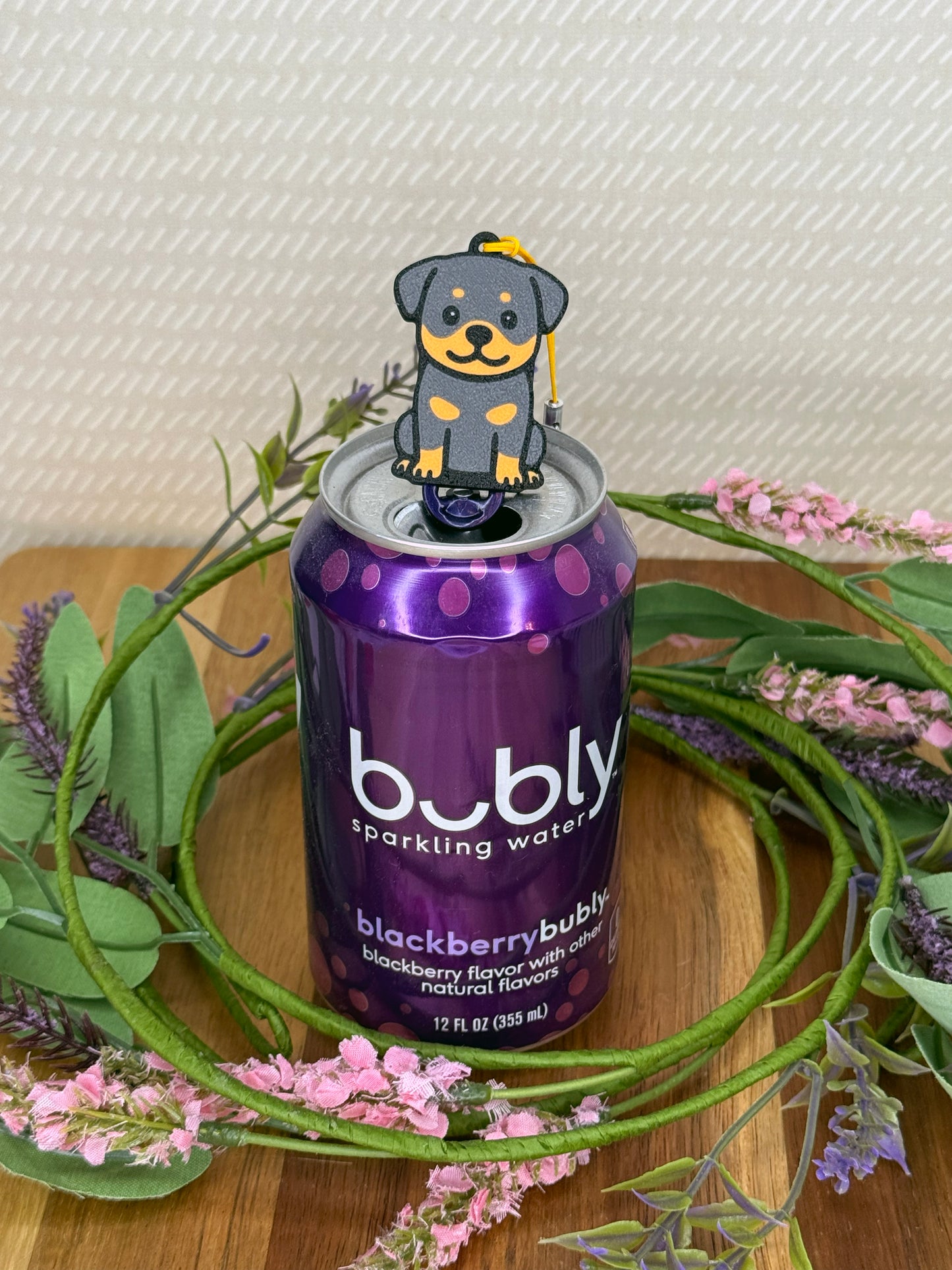 A Rottweiler design Tab Buddy rests atop the tab on a purple can of Bubly sparkling water. A wreath of flowers surrounds the can and a beige wallpaper is in background.