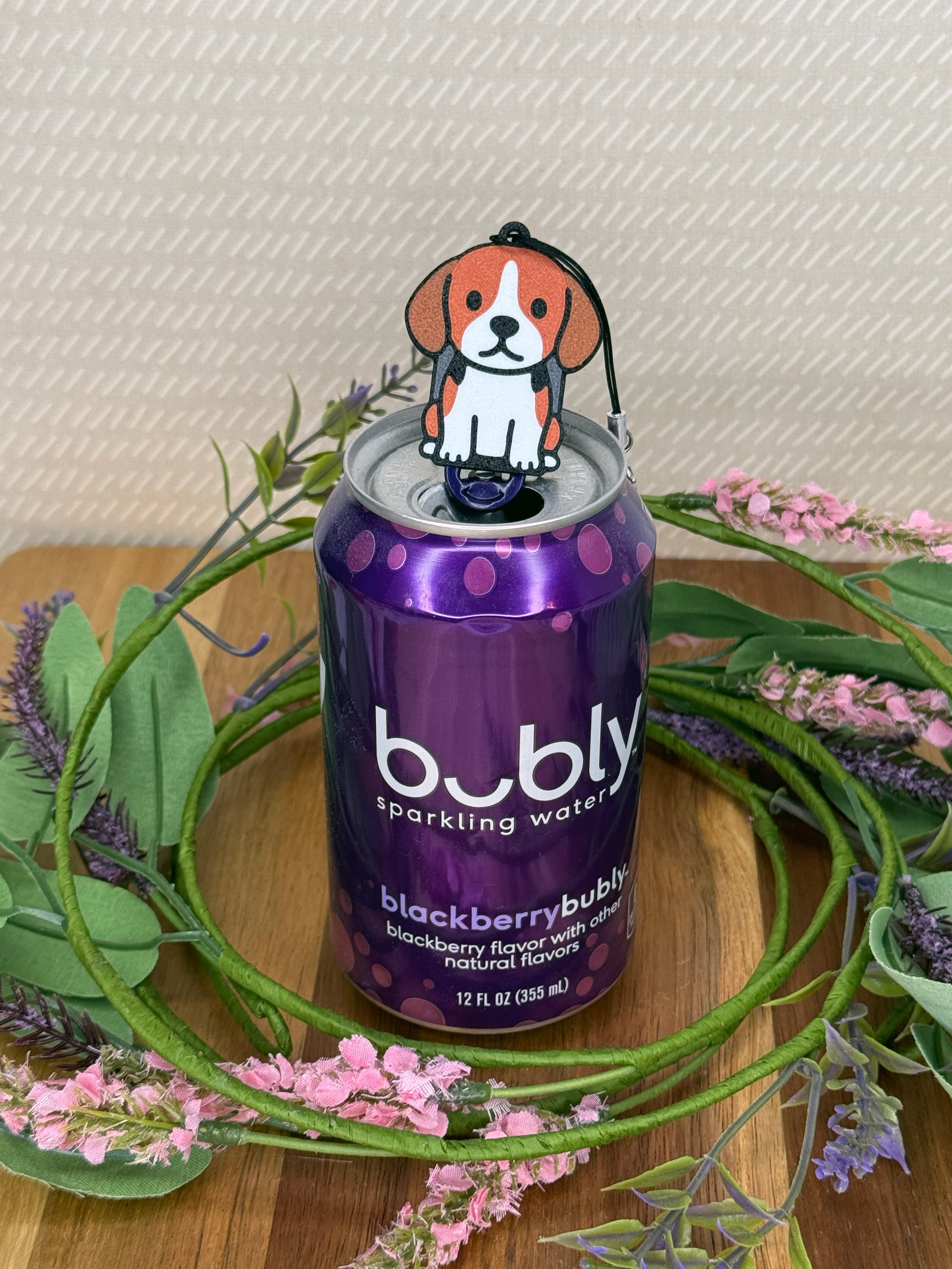 A Beagle Tab Buddy rests atop the tab on a purple can of Bubly sparkling water. A wreath of flowers surrounds the can and a beige wallpaper is in background.