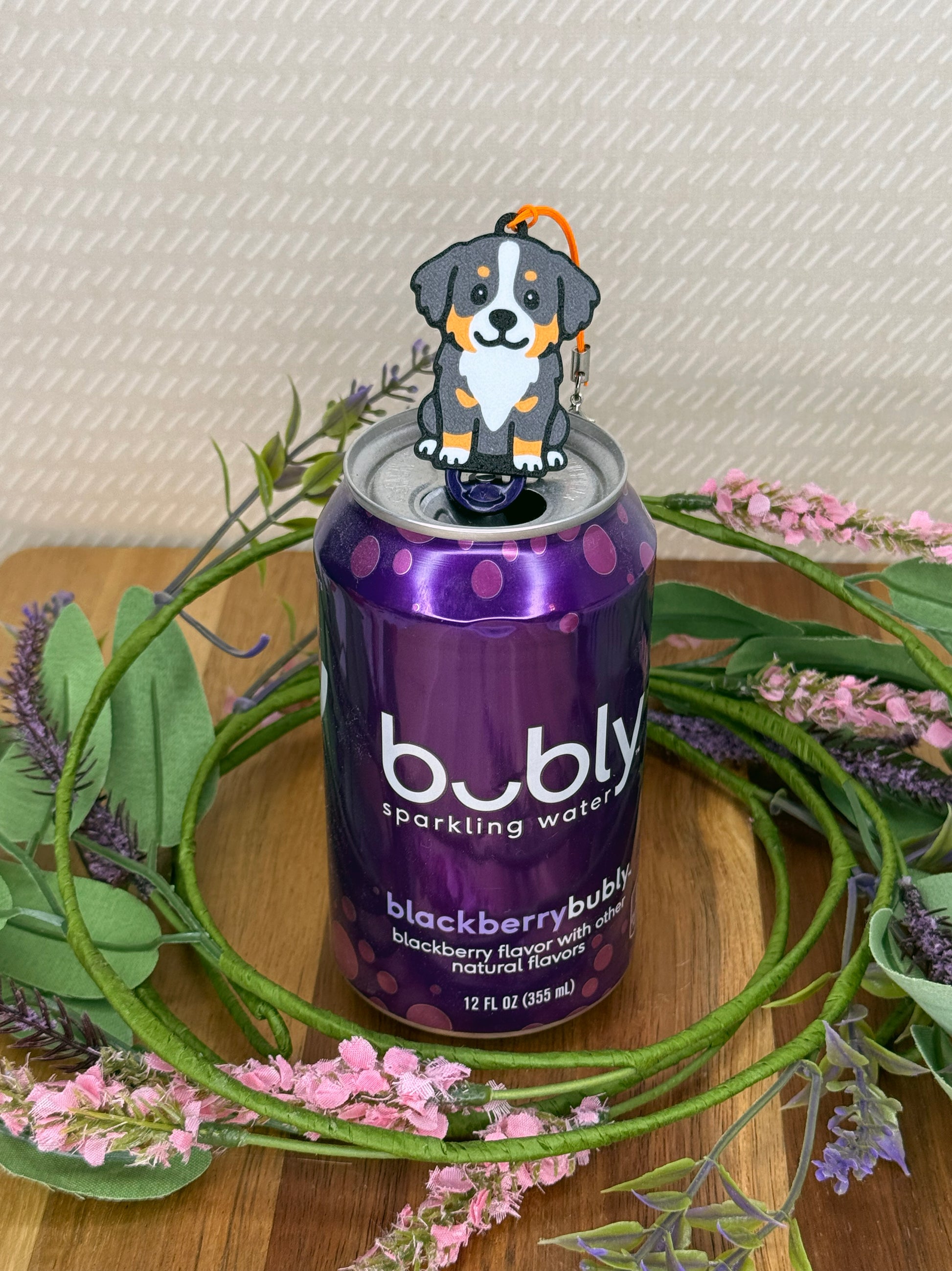 A Bernese Mountain Dog Tab Buddy rests atop the tab on a purple can of Bubly sparkling water. A wreath of flowers surrounds the can and a beige wallpaper is in background.