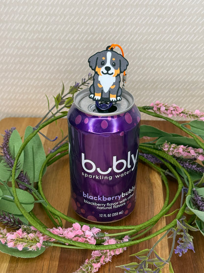 A Bernese Mountain Dog Tab Buddy rests atop the tab on a purple can of Bubly sparkling water. A wreath of flowers surrounds the can and a beige wallpaper is in background.