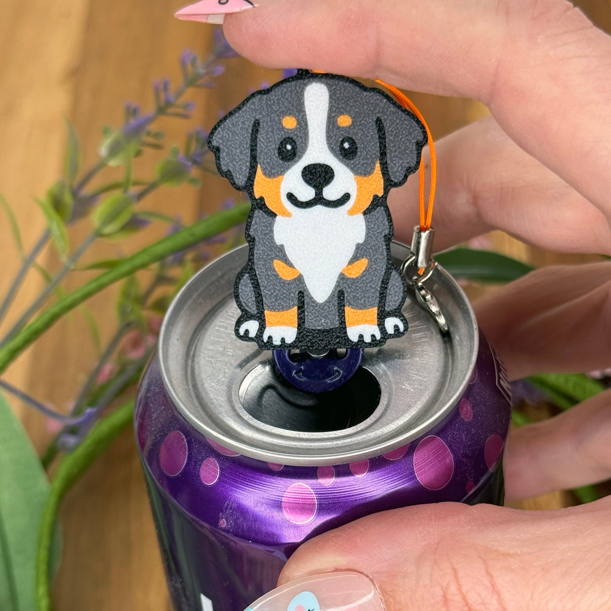 A person with long fingernails rests a finger atop a Bernese Mountain Dog design Tab Buddy after using it to open a purple can of sparkling water.