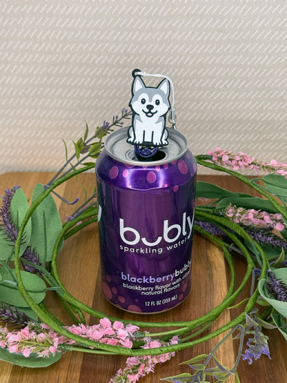 A grey and white Husky Tab Buddy rests atop the tab on a purple can of Bubly sparkling water. A wreath of flowers surrounds the can and a beige wallpaper is in background.