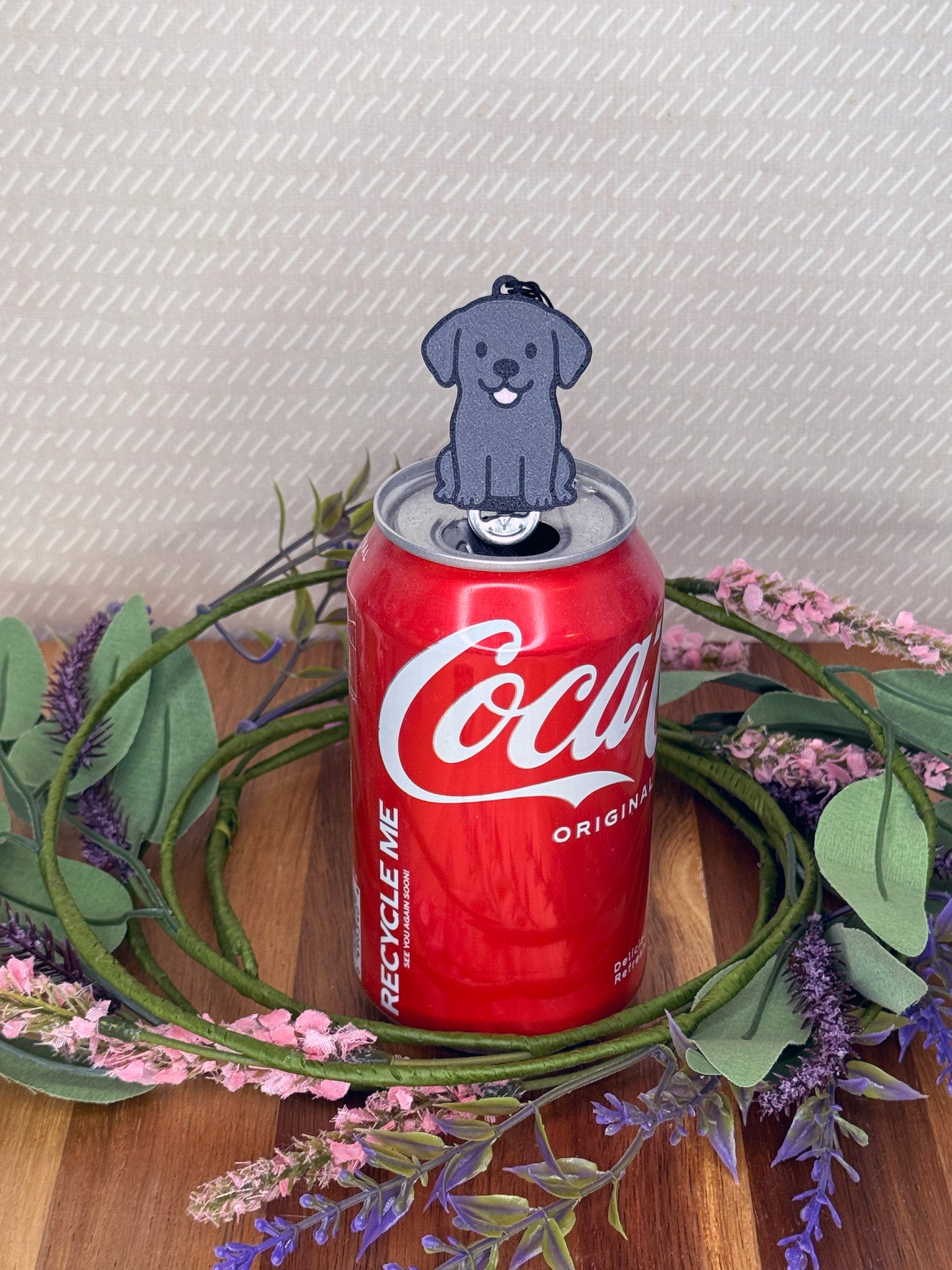 A Black Lab-inspired Tab Buddy rests atop an open can tab with a grey baackgound and floral accents.