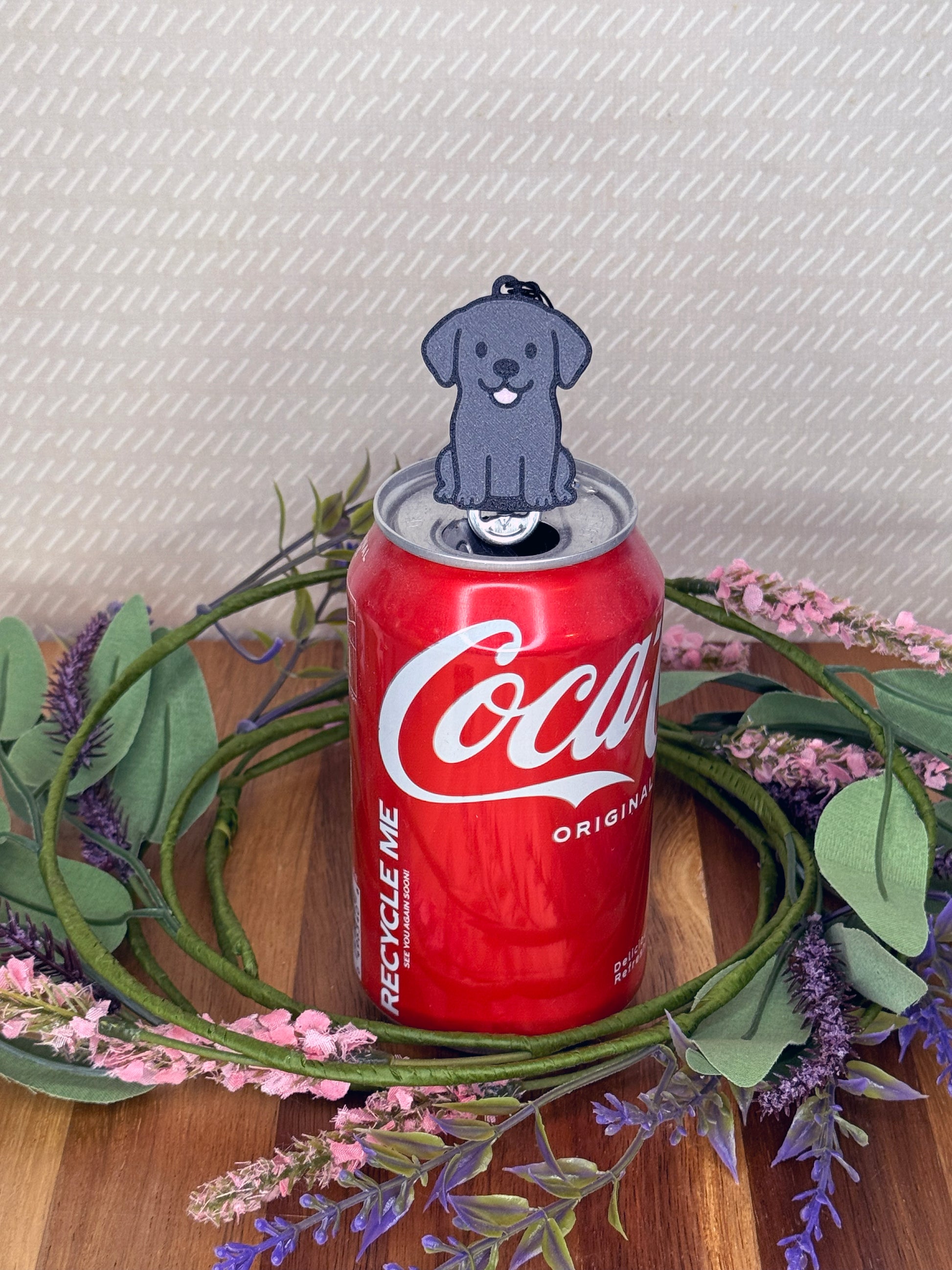 A Black Lab-inspired Tab Buddy rests atop an open can tab with a grey baackgound and floral accents.