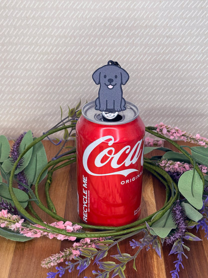 A Black Lab-inspired Tab Buddy rests atop an open can tab with a grey baackgound and floral accents.