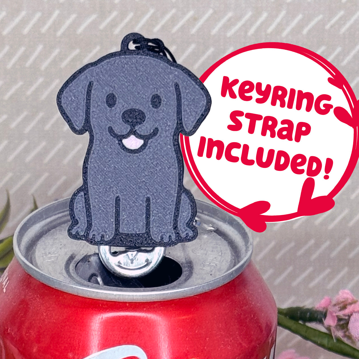 A Black Lab-inspired Tab Buddy rests atop an open can tab. Desecriptive text reads "Keyring Strap Included!"