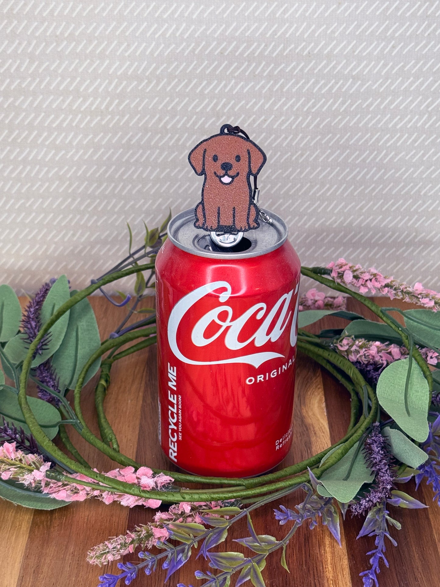 A Chocolate Lab-inspired Tab Buddy rests atop an open can tab with a grey background and floral accents.