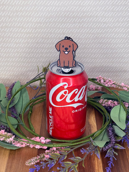A Chocolate Lab-inspired Tab Buddy rests atop an open can tab with a grey background and floral accents.