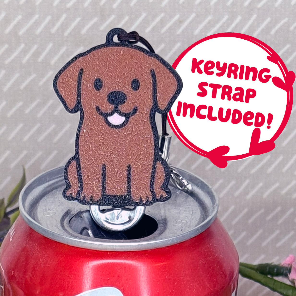 A Chocolate Lab-inspired Tab Buddy rests atop an open can tab. Desecriptive text reads "Keyring Strap Included!"