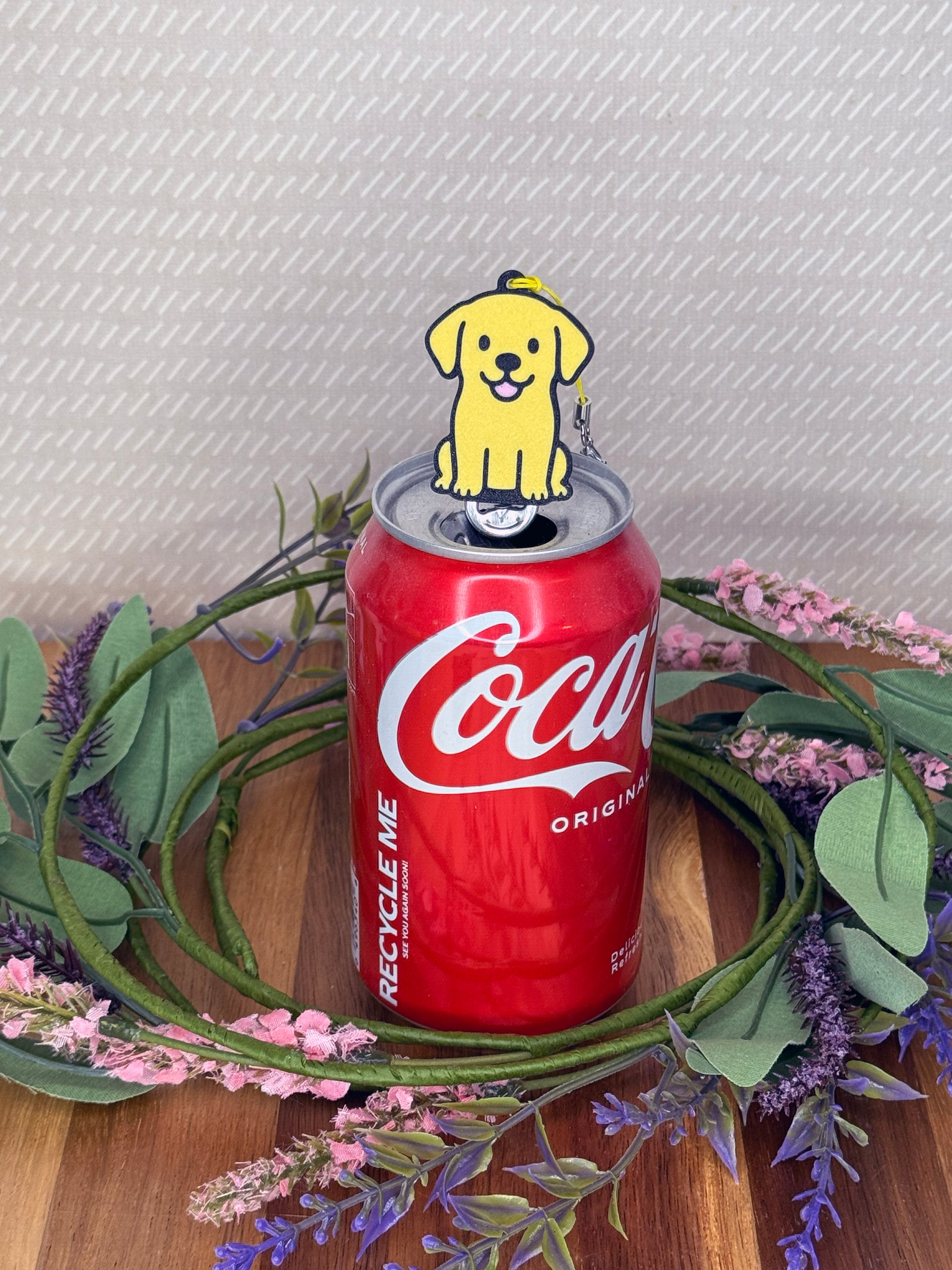 A Yellow Lab-inspired Tab Buddy rests atop an open can tab with a grey background and floral accents.