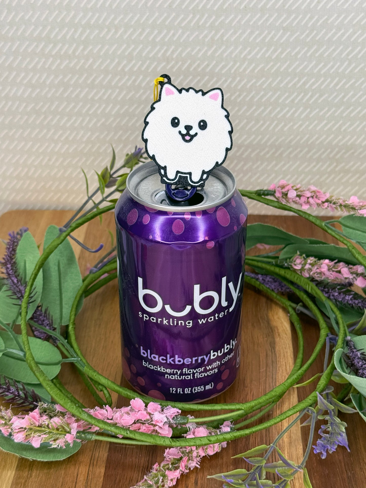 A Pomeranian design Tab Buddy rests atop the tab on a purple can of Bubly sparkling water. A wreath of flowers surrounds the can and a beige wallpaper is in background.
