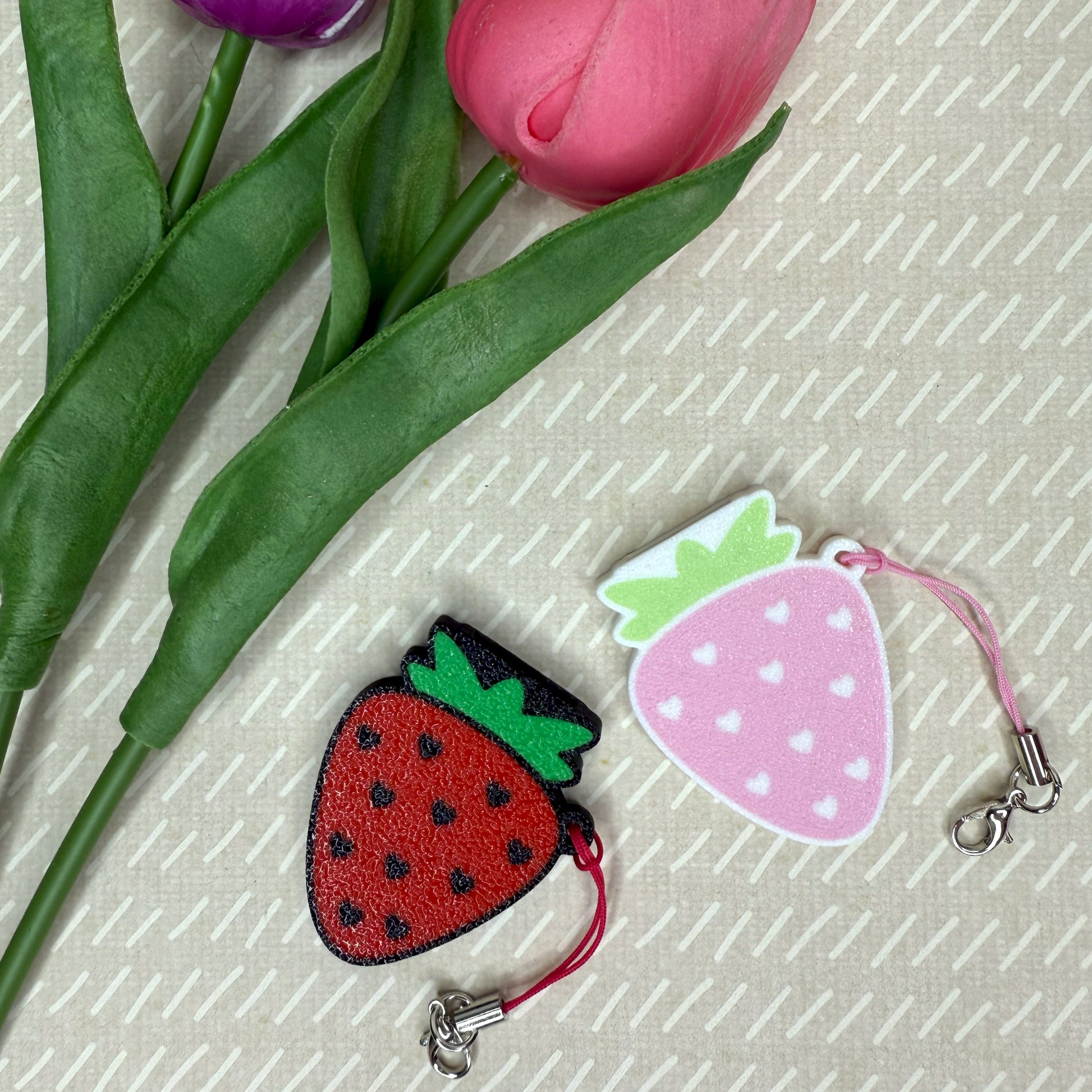 Two strawberry-shaped Tab Buddies in red and pink on a textured surface with pink tulips.