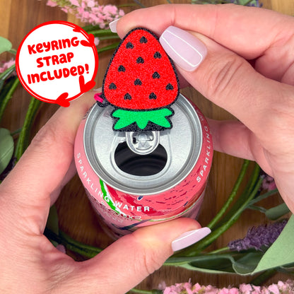 Hand holding a can of sparkling water with a red strawberry-shaped Tab Buddy with strap attached, surrounded by flowers.