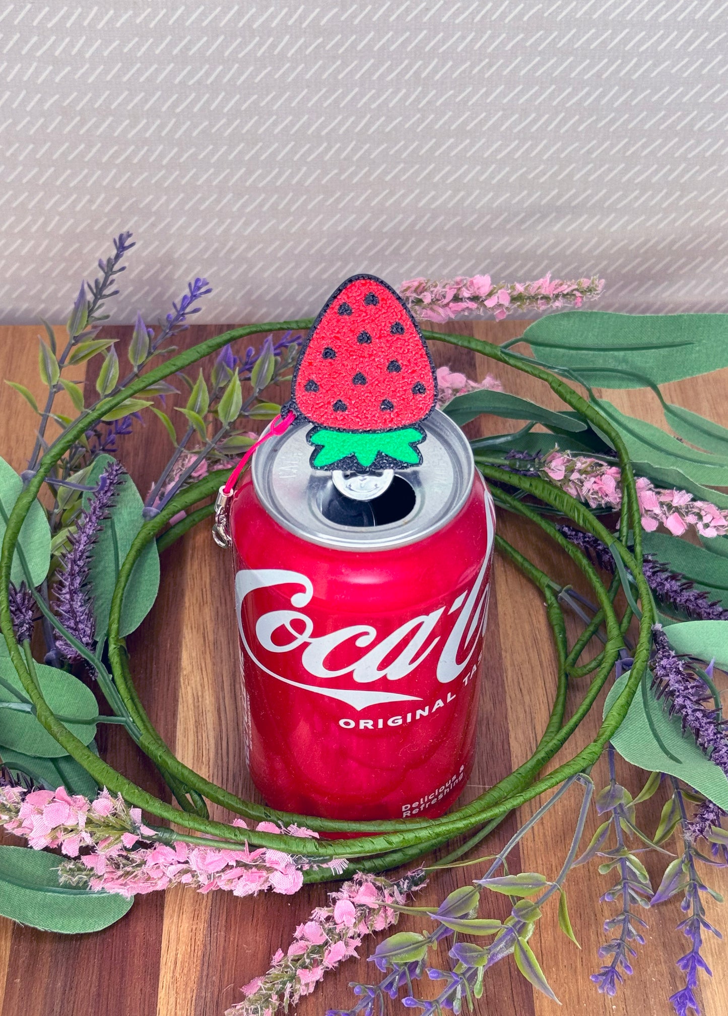 Coca-Cola can with a red strawberry Tab Buddy sitting atop a wooden surface with greenery and flowers.